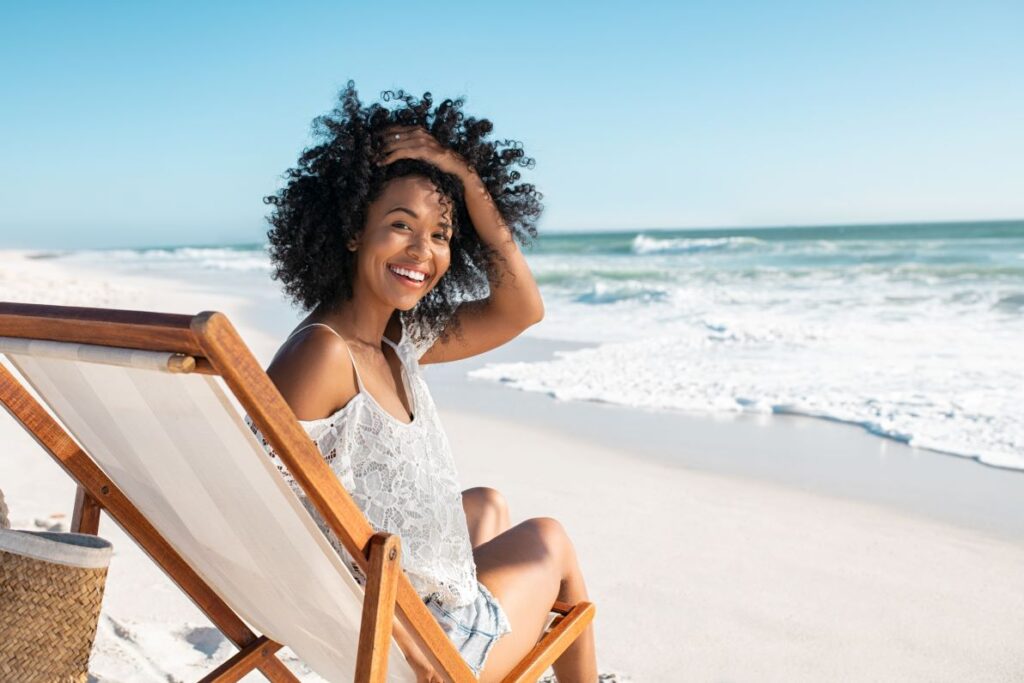A woman on a lounge chair sitting on the beach in the sun.